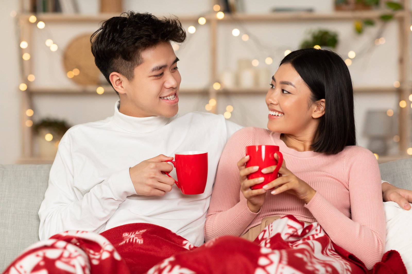 Joyful Asian Spouses Drinking Coffee Holding Mugs And Hugging Celebrating Christmas Eve Together Sitting On Sofa In Cozy Living Room At Home. Romantic Winter Date And Xmas Holidays Celebration